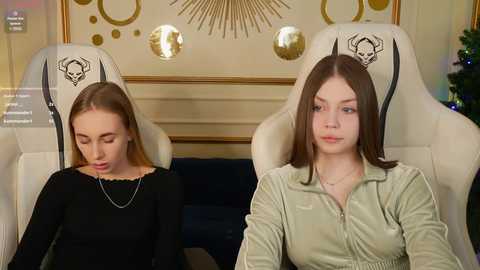 Media: Video of two young women seated in white gaming chairs with black skulls on the headrests, wearing black and green shirts, respectively, in a modern, warmly lit room with a golden, sunburst wall art and a decorated Christmas tree in the background.