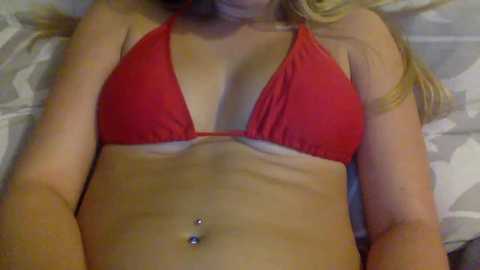 Media: Video of a fair-skinned woman with long blonde hair, lying on a bed with white and gray patterned sheets. She wears a red bikini top, revealing a pierced navel and a tan line.