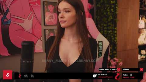 Media: Video of a young woman with long brown hair, wearing a low-cut black top, in a room with pink decor and various posters.