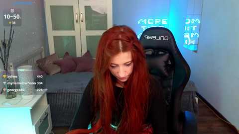 Media: Video of a young woman with long, red hair, sitting in a black gaming chair, wearing a black top, in a modern bedroom with a bed, desk, and blue lighting.