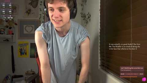 A video of a young man with short, brown hair, wearing headphones, a light blue t-shirt, and a wide smile, in a cozy, wallpapered room with framed art and a window.