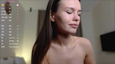 Media: A video of a nude, fair-skinned woman with long, straight brown hair, wearing minimal makeup, standing indoors, with a blurred background featuring a TV and a door.