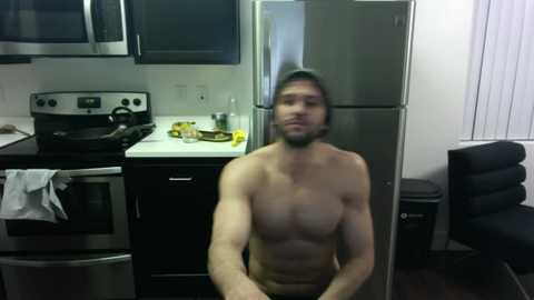 Media: A shirtless, muscular man with a beard stands in a modern kitchen with stainless steel appliances, black cabinets, and a black chair.