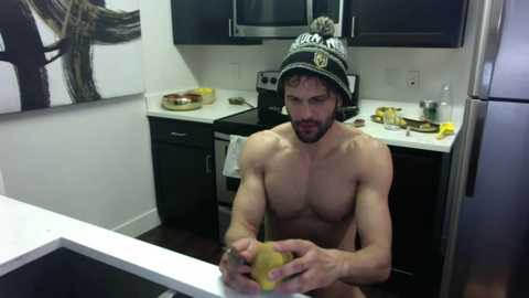 Media: Video of a shirtless, muscular man with a beard, wearing a hat, sitting in a modern kitchen, holding a banana. Background includes black cabinets, white countertops, and a stainless steel fridge.