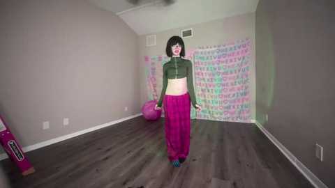 Media: A video of a young woman with a pale complexion and short black hair, wearing a green crop top, magenta pants, and blue shoes, dancing in a room with colorful, pastel-patterned wallpaper and a pink exercise ball.