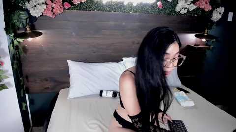 Media: Video of an Asian woman with long black hair, glasses, and black lingerie, sitting on a bed with white sheets and a wooden headboard, surrounded by floral decorations.