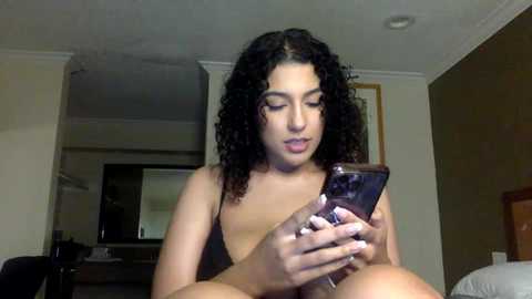 Media: Video of a young woman with curly black hair, medium skin tone, and a slender build, wearing a black bra, sitting on a bed in a hotel room, holding a smartphone.