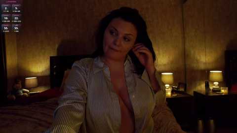 Media: Video of a middle-aged woman with dark hair, wearing a partially unbuttoned striped shirt, sitting on a bed in a dimly-lit room with two bedside lamps.