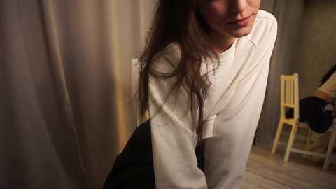 Media: Video of a young woman with long brown hair, wearing a white sweater, leaning against a wooden chair in a dimly lit room with beige curtains.