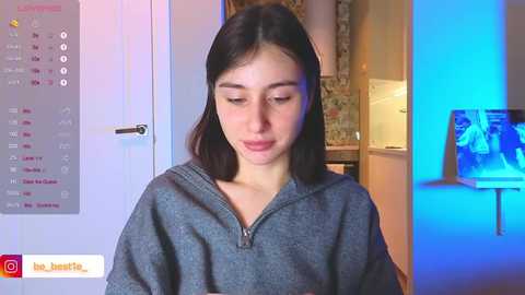 Media: Video of a young woman with straight, dark hair, wearing a gray hoodie, standing in a modern kitchen with a blue light.