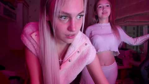 Media: Video of two young women with pale skin and long blonde hair in a dimly lit room, wearing white cropped tops, with one leaning forward, one standing behind.