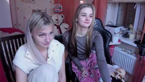 Media: Video of two young girls with blonde hair in a cozy room, one in a white sweater, the other in a brown top and patterned skirt, surrounded by stuffed toys and a radiator.