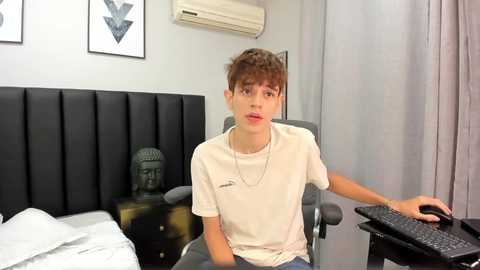 Media: Video of a young Caucasian male with short brown hair, wearing a beige t-shirt, sitting on a grey chair in a modern bedroom.