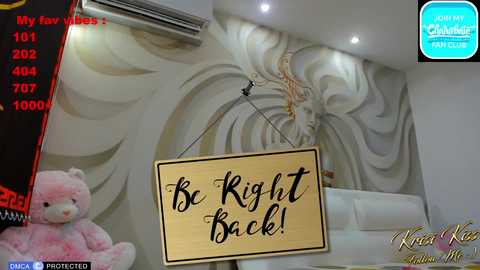 A video of a modern living room with a large, gold-framed sign reading \"Be Right Back!\" hanging from the ceiling. A plush teddy bear sits on a white couch, with a decorative wall mural in the background.