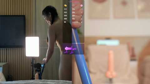 Media: Video of a nude woman with dark hair, holding a vibrator, standing next to a bed in a modern bedroom with wooden paneling and a lit lamp.