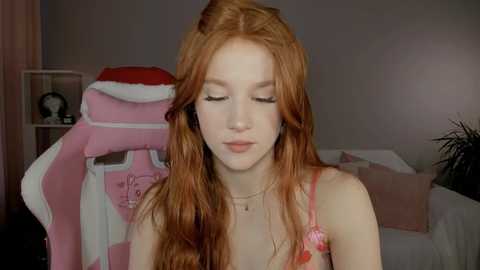 Media: Video of a fair-skinned, red-haired young woman with long, wavy hair, wearing a pink lace bra and a delicate necklace, sitting in a cozy, dimly lit bedroom with a gaming chair and a plant in the background.