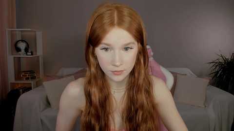 Media: Video of a pale-skinned, long-haired redhead woman with soft features, sitting on a white couch, wearing a pink top. Background includes a bookshelf with decorative items and a plant.