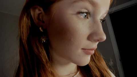 Media: Video of a young Caucasian woman with fair skin, long auburn hair, and freckles, looking to the side in a dimly lit room. She wears a necklace and earrings, her expression is contemplative.