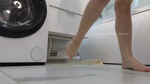 A video of a woman in a white, modern bathroom, barefoot, mopping the floor. The background shows a black washer and dryer, a white sink, and a window.