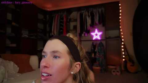 Video of a blonde woman with a black headband, wearing a black shirt, in a dimly lit bedroom with a star-shaped neon light, clothes, and guitar visible.