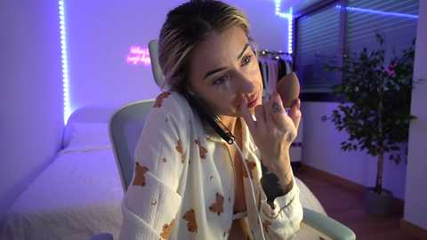 Media: Video of a young woman with light skin, blonde hair, and a white hoodie with brown stains, wearing headphones, sucking a lollipop in a dimly lit room with blue LED strip lights, a white bed, and a potted plant.