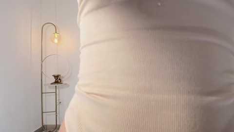 Media: Video of a woman's bare abdomen, wearing a tight, ribbed white tank top. Background includes a modern, minimalist lamp with a curved arm and a small stool. The scene is lit softly with a warm, ambient glow.