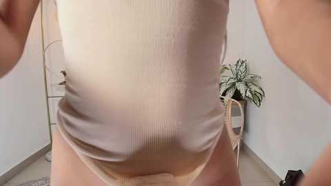 Media: Video of a person in a beige ribbed tank top and beige underwear, standing in a minimalistic room with a white wall and potted plant in the background.