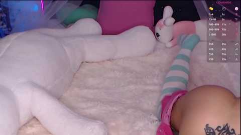 Media: Video of a white, plush bunny lying on a pink, fluffy bedspread, with a person in a pink top and striped socks visible in the foreground. Background shows a pink and green pillow.