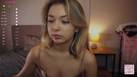 Media: Video of a young woman with light skin, blonde hair, and a slim figure, wearing a beige camisole, in a dimly lit bedroom with a bedside lamp and a bed with a pink blanket.