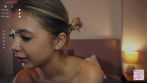 Media: A video of a light-skinned woman with blonde hair tied back, wearing a nose ring and makeup, captured in a dimly lit bedroom with a warm, ambient glow.