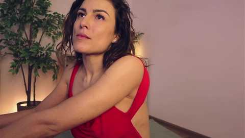 Media: Video of a young woman with wet, wavy dark hair, wearing a red sleeveless dress, standing indoors against a pale pink wall, with a potted plant and soft lighting in the background.