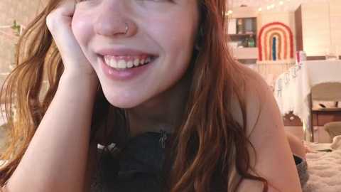 Media: A close-up video of a smiling young woman with long, wavy brown hair, wearing a black top, lying on a plush carpet in a cozy, brightly lit room with a rainbow poster in the background.