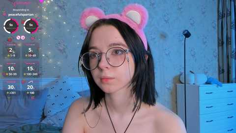 Media: Video of a young woman with shoulder-length black hair, wearing a pink bear headband, glasses, and a black necklace, standing in a bedroom with a white dresser and floral wallpaper.