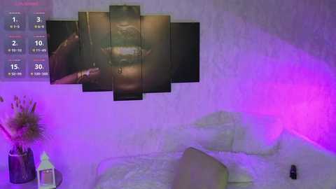 Media: Video of a dimly-lit bedroom with a white bed, lavender and purple lighting, a black framed video of a woman's face, a glass vase with dried flowers, and a white candle on a nightstand.