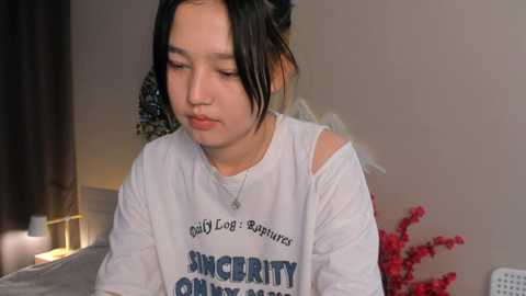 Media: Video of a young Asian woman with medium-length black hair, wearing a white long-sleeve shirt with \"Sincerity\" text, sitting indoors, looking contemplative, with a beige wall background.