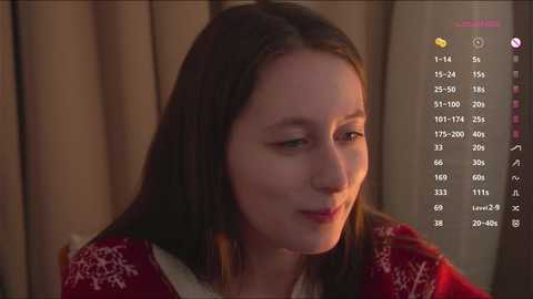 A video of a smiling young woman with long brown hair, wearing a red and white patterned sweater, indoors against a beige curtain background.