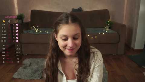 Media: Video of a smiling young woman with long, wavy brown hair, wearing a white blouse, sitting on a gray rug in a cozy living room with a brown couch, string lights, and wooden floor.