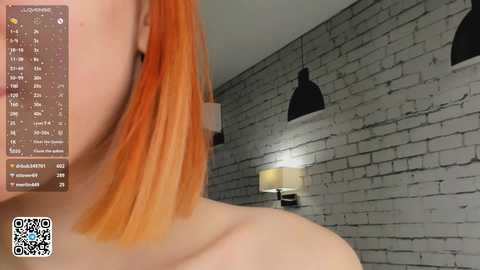 Media: A video of a young woman with short, vibrant orange hair, wearing a strapless top, standing in a modern room with a gray brick wall, black pendant lights, and a lit table lamp.