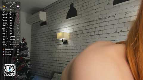 Media: A video of a woman with red hair, partially visible, standing in a modern living room with a white brick wall, black pendant light, and a Christmas tree on the left.