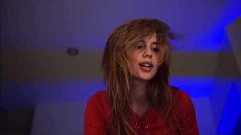 Media: Video of a young woman with long, messy blonde hair, wearing a red top, singing passionately in a dimly lit room with blue lighting.