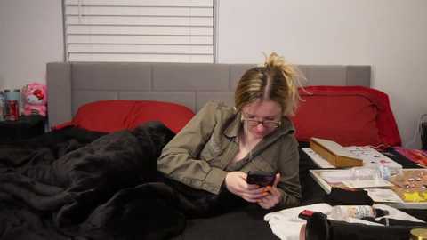 Media: A video of a blonde woman in glasses, wearing a green jacket, lying on a bed with red and black sheets, reading a book, surrounded by scattered objects and a partially visible window with white blinds.