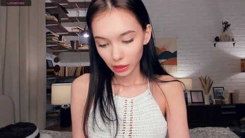 Video of an Asian woman with long black hair, wearing a light gray crochet top, in a cozy, modern living room with white brick wall, shelves, and a table lamp.