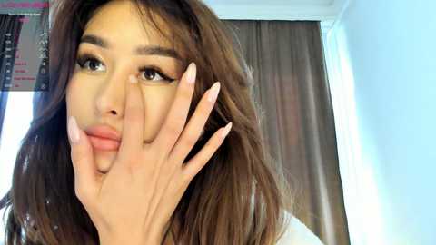 Media: Video of a young woman with long brown hair, fair skin, and full lips, playfully covering her eyes with her fingers. Background features a light blue wall and brown curtains.