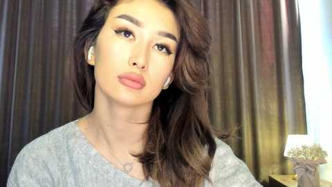 Media: Video of an Asian woman with long, wavy brown hair, wearing a light gray sweater, posing against dark, textured curtains. She has a neutral expression, light makeup, and small hoop earrings.