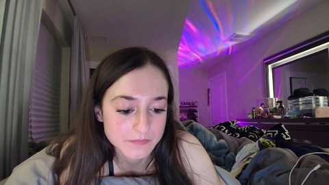 Media: Video of a young woman with long brown hair, wearing a black top, lying on a messy bed, under purple LED strip lights. Background shows a cluttered room with a dresser and mirror.