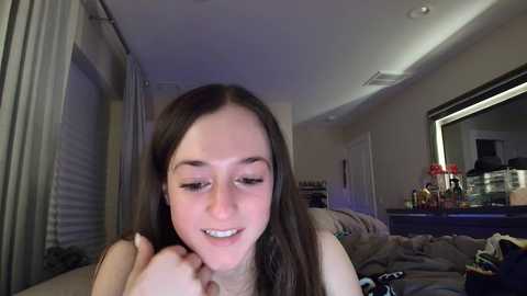 Media: Video of a young woman with long brown hair, smiling, in a dimly lit bedroom. She's lying on a messy bed, with a dresser and mirror in the background.