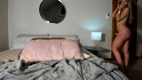 Media: Video of a nude, slender blonde woman with long hair taking a selfie in a dimly lit bedroom. The bed has a pink blanket and gray throw, a round mirror, and a nightstand with a lamp.