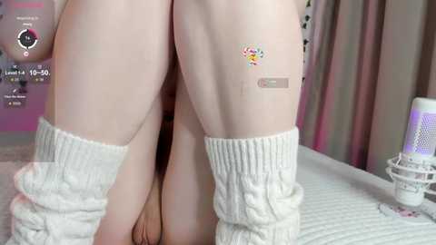 Media: Video of a light-skinned person wearing white knit leg warmers, seated with legs bent and knees together, revealing their vulva. Background features a bed with soft, pastel-colored bedding and a decorative lamp.