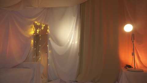 Media: Video of a softly lit, cozy room with sheer white drapes, fairy lights, a warm orange lamp, and a round table, creating a serene, romantic ambiance.