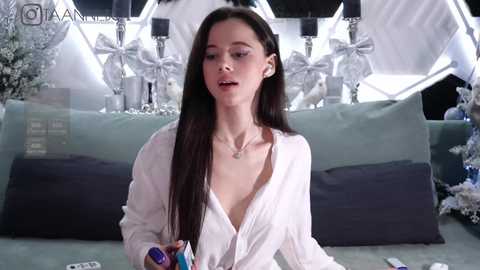 Media: Video of a slender, young Caucasian woman with long dark hair, wearing a loose white blouse, sitting on a grey sofa in a modern, minimalist room.